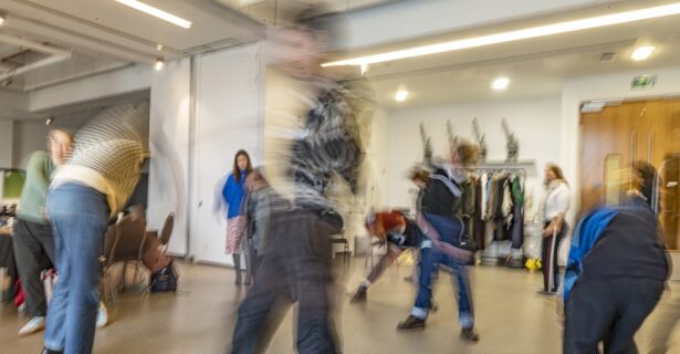 A long-exposure photograph taken in a brightly lit, modern studio space at M Shed. The slow shutter speed creates a sense of high energy and motion, with several figures appearing as blurred, ghost-like streaks as they move across the room. In the background, a person in a blue top stands still, providing a sharp point of contrast to the kinetic energy of the dancers.