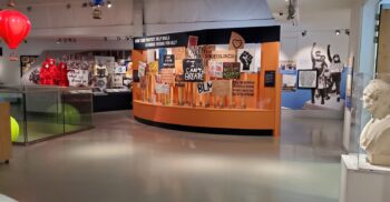 An interior view of a museum gallery featuring a large, curved orange display stand filled with hand-painted protest placards. A sign above the display reads, 