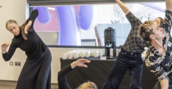 A clear, still photograph of four people engaged in a contemporary dance workshop. One person is seated on the floor in a cross-legged position, leaning to the side with an arched arm. Three other dancers stand behind them, also captured in mid-movement with expressive, reaching gestures. The setting is a minimalist room with a large window and a table with refreshments in the background.